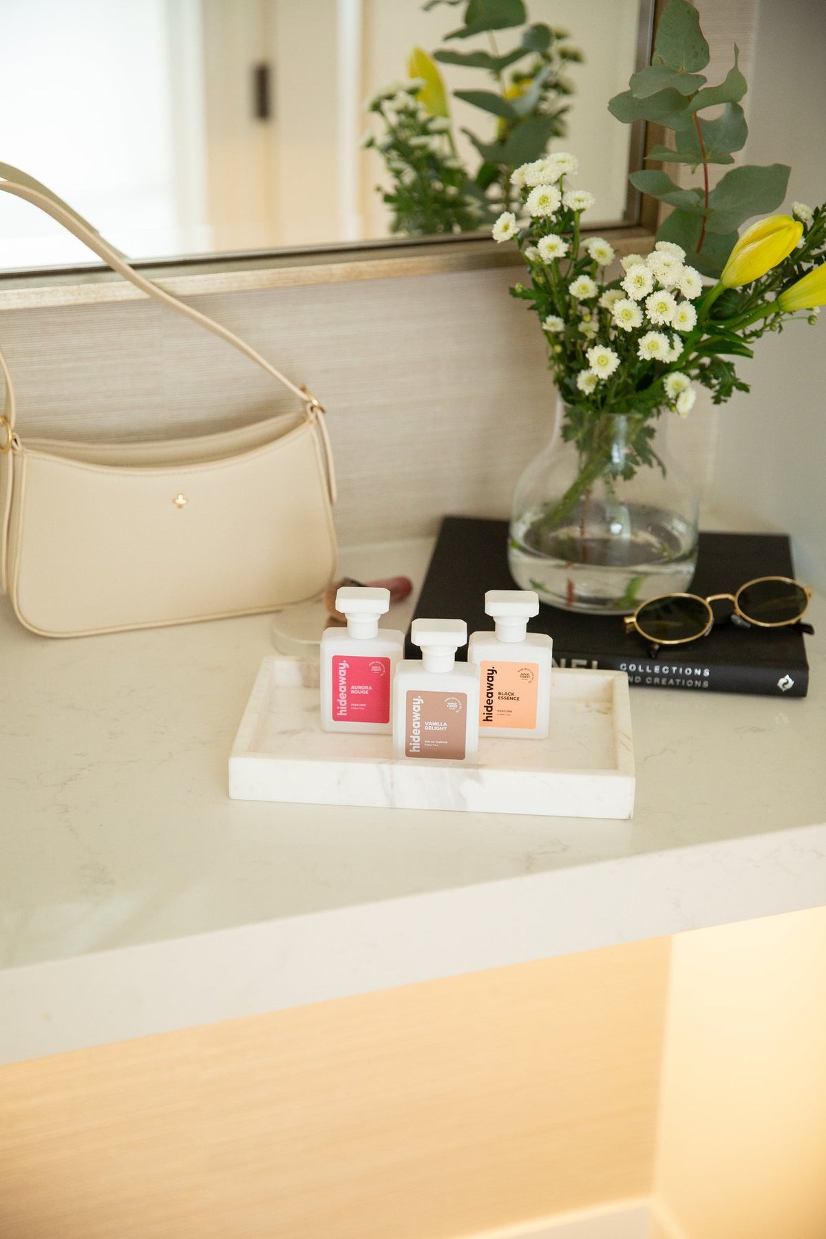 Three hideaway. perfumes on a vanity tray beside a handbag and flowers