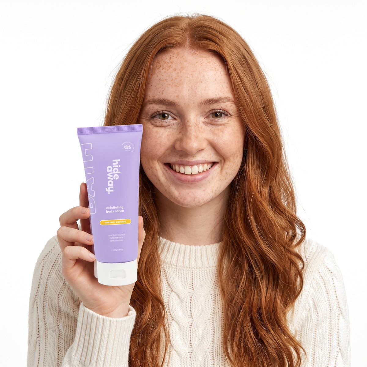 A smiling woman holding Hideaway. Pineapple Coconut exfoliating body scrub